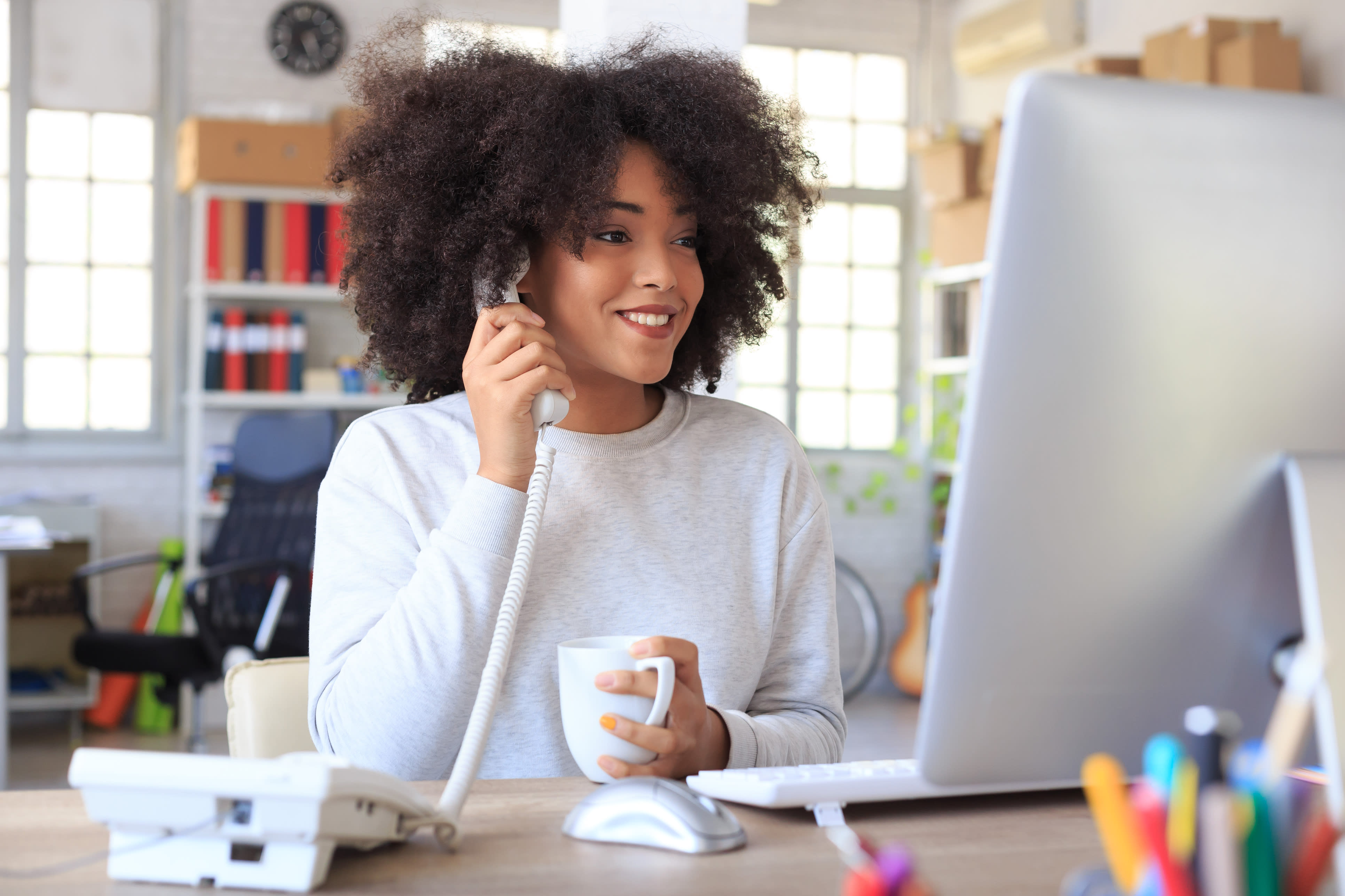 Woman works on a computer and talks on a landline corded phone at work.